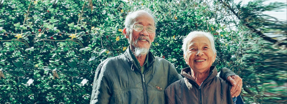 Older man and woman couple smile at the camera standing in front of trees and vines