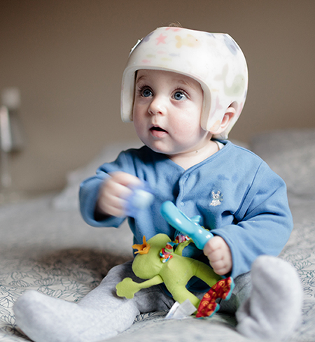 A photo of an infant who is wearing a therapy helmet while sitting on a bed and playing with toys.