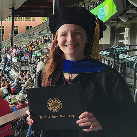 A photo of Betts Peters smiling in cap-and-gown holding her Ph.D. degree from Portland State University at her graduation.