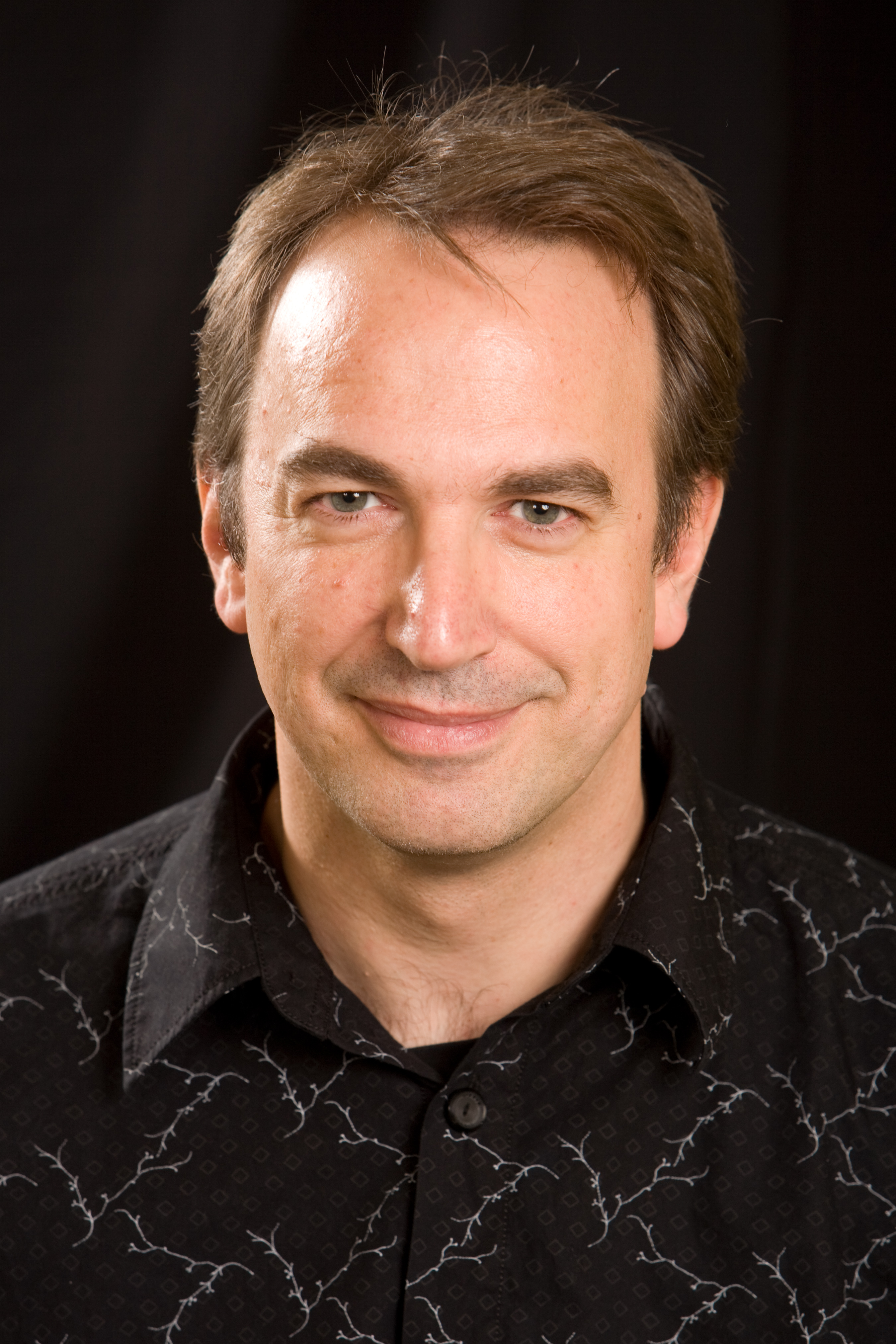 headshot of white man with brown hair wearing a black shirt smirking in front of a black background