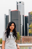 Dr. Leena Kadam has long dark hair and is wearing a short sleeve gray shirt. She is standing in front of several tall buildings.