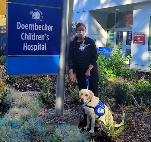 Casey the dog sits next to a sign for Doernbecher Children’s Hospital while handler Nikki Wiggins holds her leash.