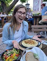 A woman smiling with a newly served plate of food at an outdoor dining table.