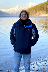 A man smiling while standing in front of a lake with mountains in the background.