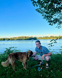 A man crouching down smiling next to his dog in front of a lake.