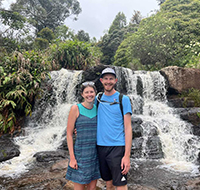 A woman and a man smiling in front of a river with a small waterfall.