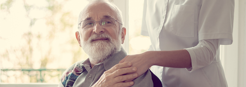 A health care provider standing behind a sitting elderly man and placing their hand on his shoulder, as he reaches back and puts his hand on top of theirs.