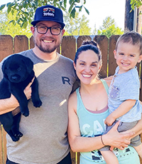 A man holding a black puppy next to a woman holding a baby, all smiling outside in front of a wooden fence.