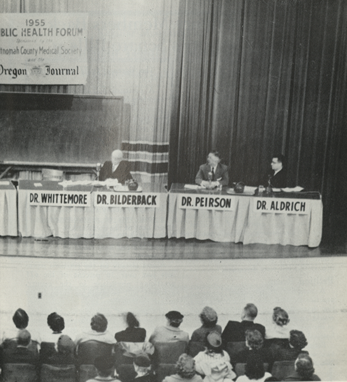 Photo of physicians on a stage with an audience. Poster in the back of the stage reads "1955 Public Health Forum. Multnomah County Medical Society. Oregon Journal"