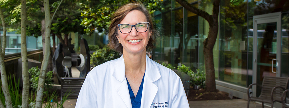 Dr. Alison Edelman stands on a wide balcony outside the OHSU Center for Women’s Health. She wears a white doctor’s coat, and smiles broadly.