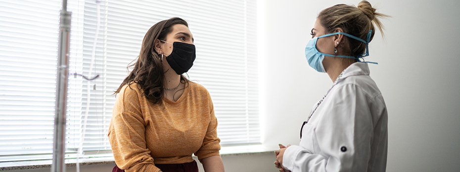 A woman sitting down talking to a female doctor standing in front of her, both wearing PPE facemasks.