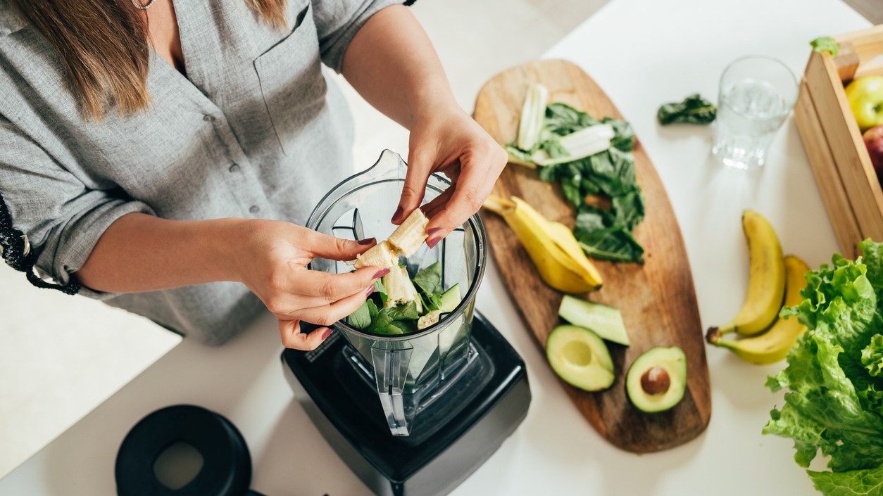 A person adds greens, avocado, and banana to a blender
