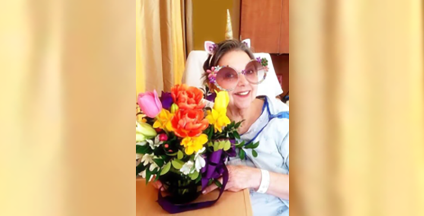 A patient sits in a hospital room. She is smiling and holding a basket of flowers.
