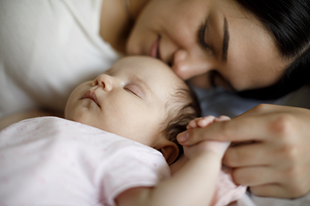 A woman smiles and rests as her sleeping baby holds her finger.