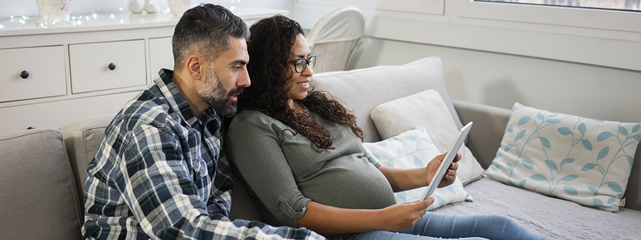 An expecting couple sits together on a couch, attending a virtual childbirth class on their tablet.