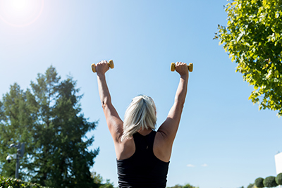 An older person is exercising outside. The sun is shining and they are framed by trees. They face away and hold two small weights above their head.