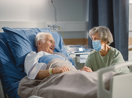 An elderly man in a hospital bed smiles at his visiting partner.