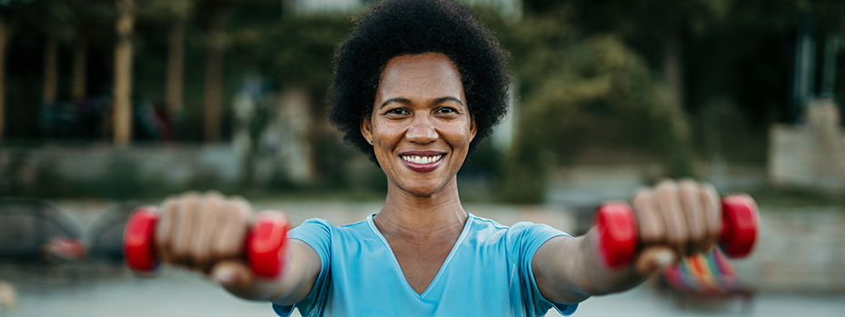 A woman is exercising outside. Her arms are stretched out toward the viewer. She is holding two small red weights. She is smiling