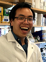 A man smiling in a white lab coat in a research lab.