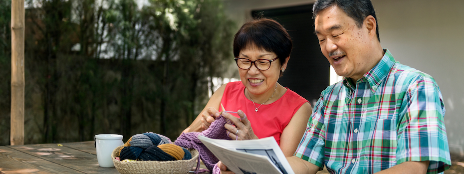 A senior couple sitting together outside, reading and knitting.