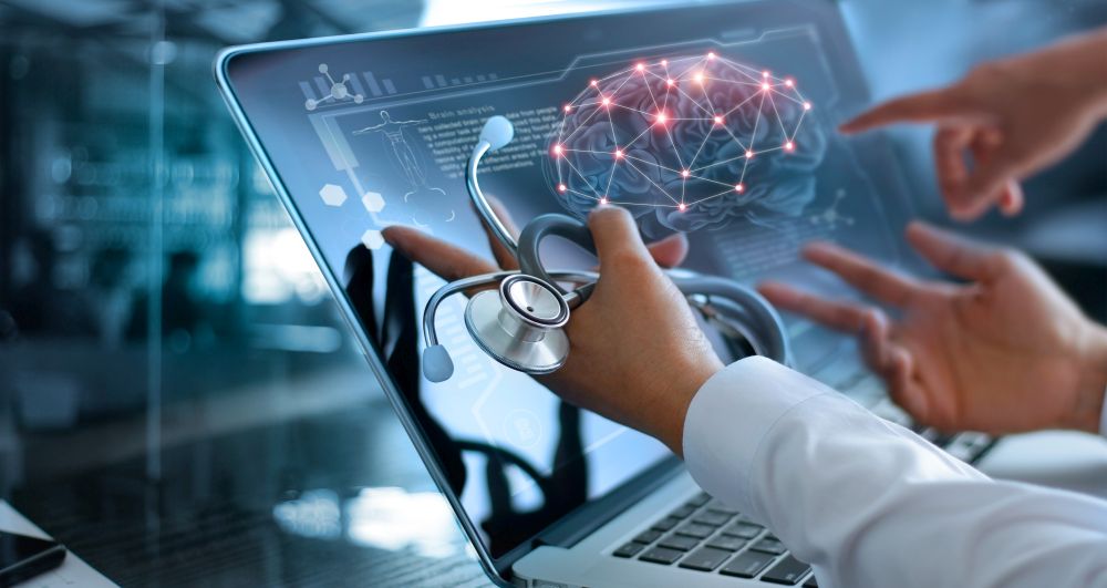 Hands from three different people pointing at a brain network diagram on a laptop screen. One hand is holding a stethoscope and the arm has a white lab coat sleeve.