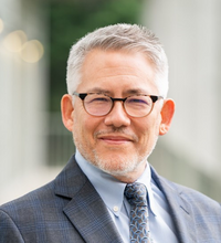 Man smiling slightly, wearing a gray suit and glasses