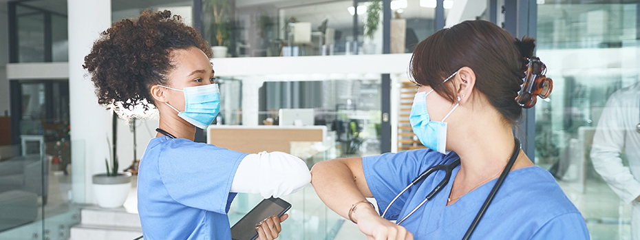 Two nurses wearing masks take a moment to smile and bump elbows.