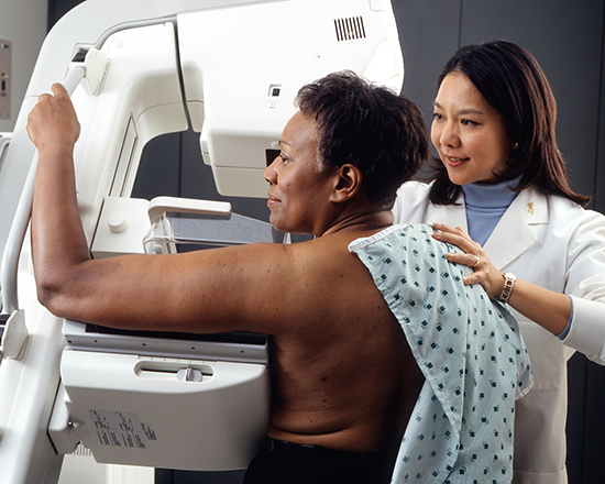 A patient is getting a mammogram. She stands in front of the mammogram machine, looking left. She is grasping a handle on the machine with one arm. A technologist is helping her. The technologist stands on the right. She is guiding the patient into the proper position by gently touching her back.
