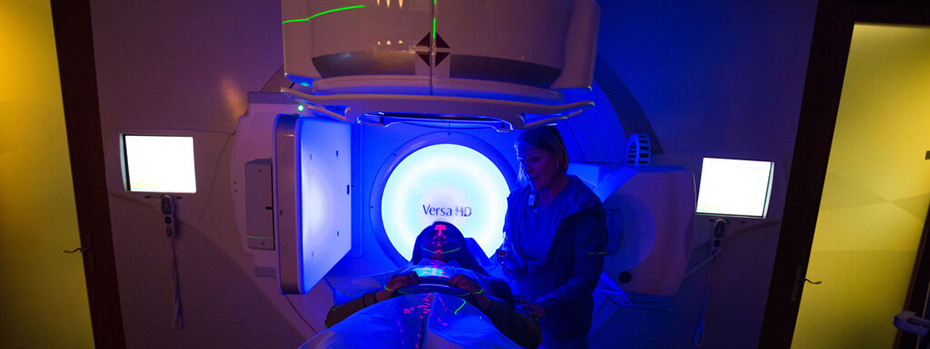 A patient is lying on the bed of a radiation therapy machine. A radiation therapist stands next to her. The room is dark but the people’s faces are illuminated by light.