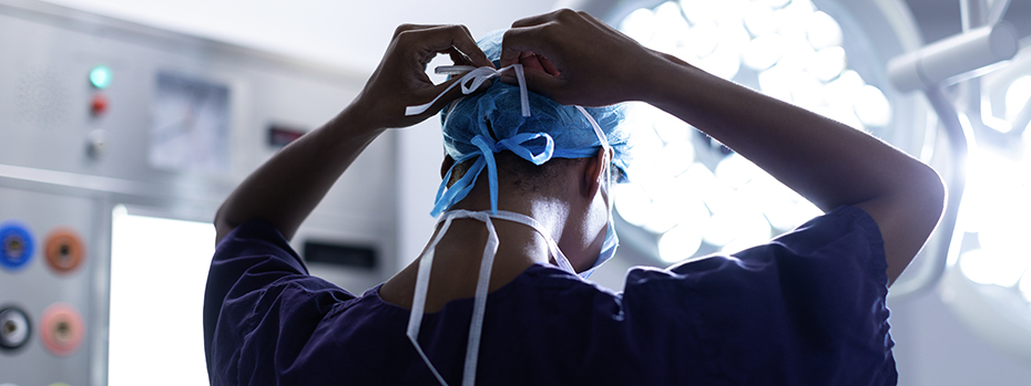 A doctor is putting on a mask in a radiation therapy room. They are facing away from the camera and adjusting the straps of the mask. The doctor is silhouetted by a powerful lamp.