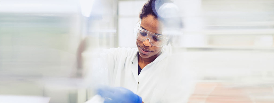 A researcher is working in a laboratory. She wears a white lab coat, protective glasses and blue gloves. She looks intently at her work.