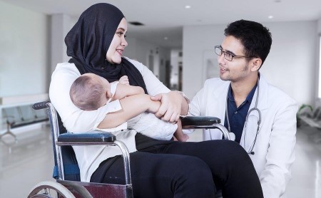 Woman with baby in arms sitting in wheelchair with male doctor kneeling beside