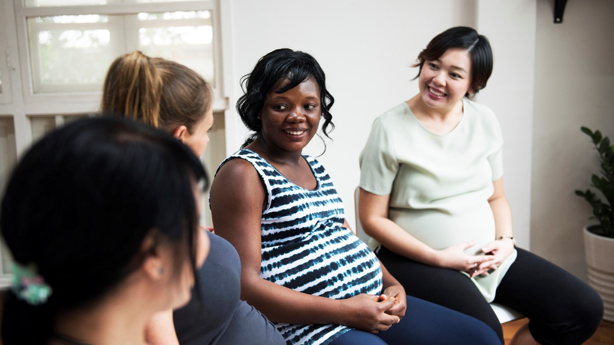 A group of pregnant people sit and talk with one another, smiling.
