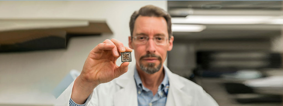 A researcher holds a computer chip in his right hand, grasping it gently with thumb and fingers. The chip is the size of a big postage stamp. The chip is in focus. The researcher’s face and body are in the background.