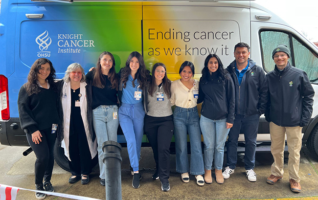 A group of 9 people is standing in front of the Knight Cancer Institute mobile van. They are happy and smiling. The van is painted blue, green and white with the tagline “Ending cancer as we know it.”