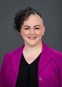 White woman with short curly black hair on half her head and bold on the other half smiling while wearing a purple blazer and black v neck shirt