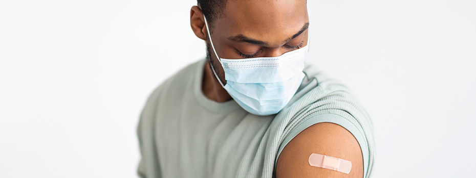 A person wearing a face mask and a T-shirt stands with one sleeve rolled up to show a Band-Aid where they were vaccinated.