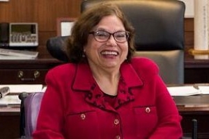 Judy Heumann sits in an office, smiling in a red shirt.