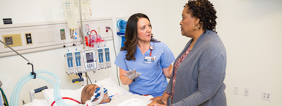 An OHSU nurse shares information with a caregiver at a patient’s bedside. The patient is breathing through a tube.
