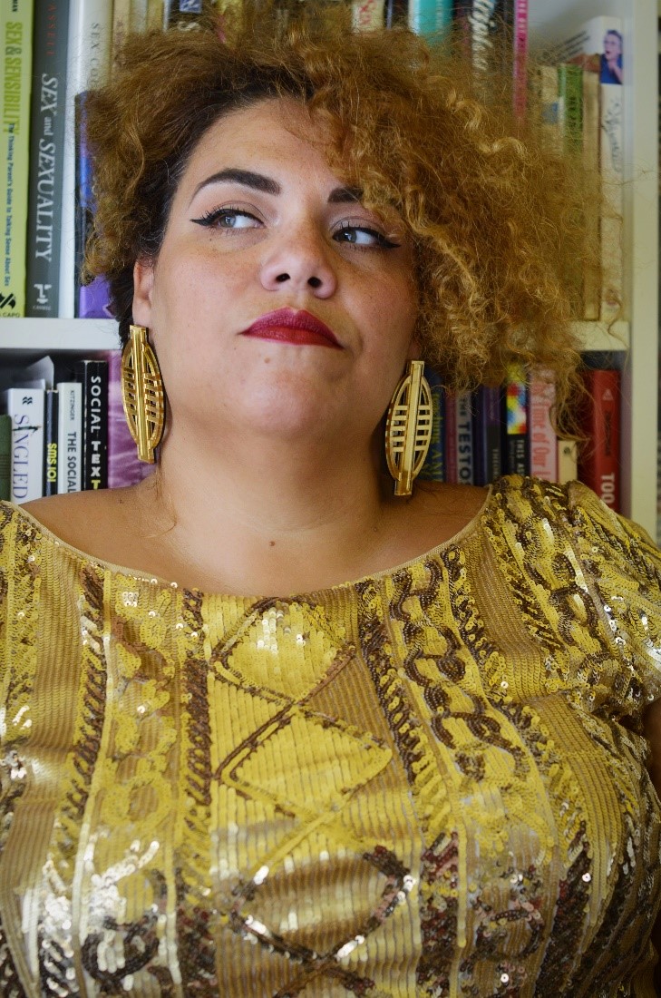 Bianca is a tall fat light skinned disabled queer AfraLatina who is in a gold dress with gold beaded geometric designs and looking to the left side of the room Her hair is up and her blondish brown curls cascade around her head. She has on red lipstick and large round gold earrings. She stands in front of a full bookcase with a serious look on her face.