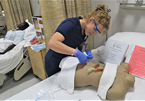 A nursing student leans over a mannequin while inserting an IV.