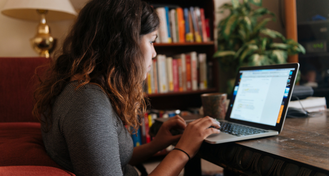 A student works on her laptop while at home. A bookcase and plant is visible in the background.