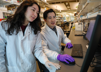 Two students in lab coats and gloves work on a computer in a lab.