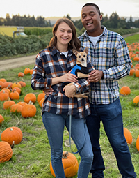 A man and a woman standing and smiling at a pumpkin patch, with the woman holding a small dog.