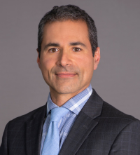 Headshot of man with blue shirt and tie, black jacket and dark short hair