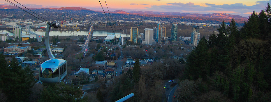 An aerial tram moves up a cable with a sunrise view of buildings and hills behind it.