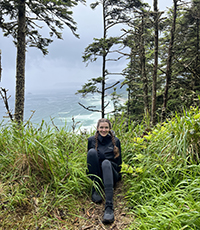 A woman sitting on the ground on a hiking trail with the ocean behind her.