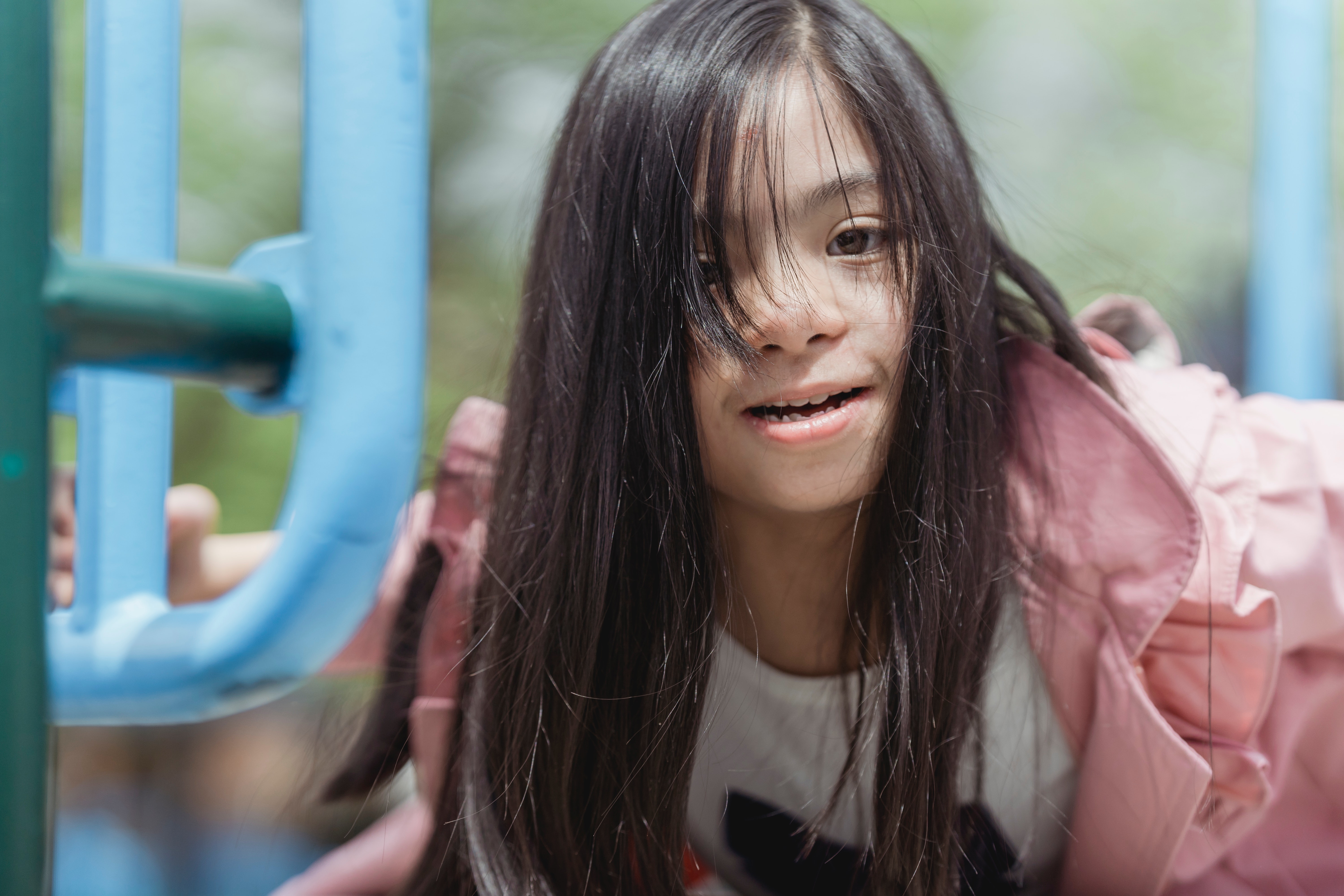 A child with long dark hair, medium-tan skin, and a pink jacket happily plays on playground equipment.
