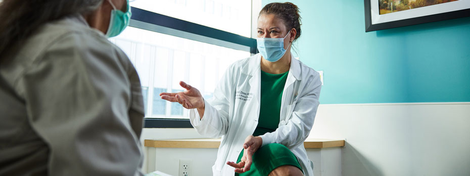 A doctor reassures a patient in a medical exam room. She wears a lab coat and gestures with her hand as she explains how treatment will help the patient’s condition.