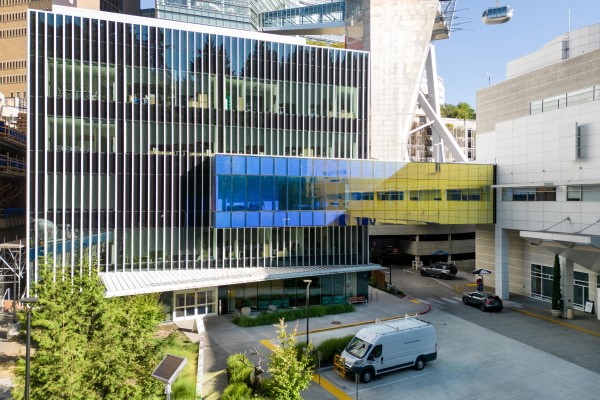 Building with several stories and all glass facing the patient drop off area and a walking bridge connecting to the neighboring building.